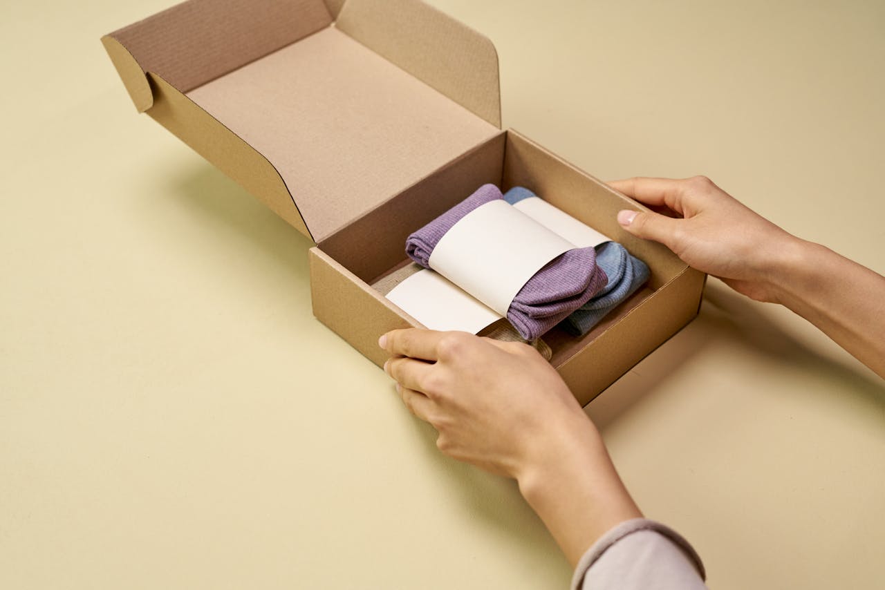 A person carefully places folded socks into a cardboard packaging box on a beige background.